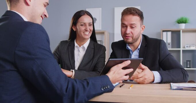 Close up of professional male real estate manager sitting at desk with couple of business people, showing alternatives at tablet screen. Cheerful man and woman exploring options for purchase or rent.
