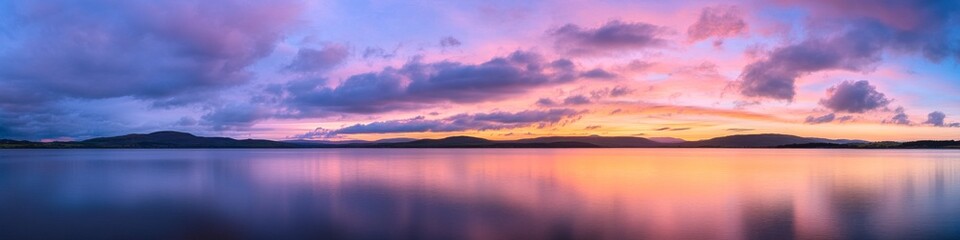 Obraz premium majestic sunset over calm waters, sweeping cloudy sky, double horizon effect, subtle warm and cool tones, distant shore vegetation, panoramic nature photograph
