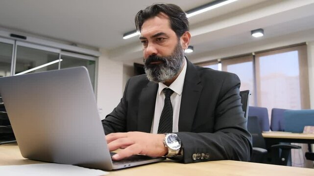 A middle-aged, dark-haired businessman in a suit is happily working at his desk in the office, using his computer