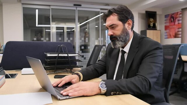A middle-aged, dark-haired, bearded businessman is at his desk in his office, experiencing tense moments during a video meeting on his computer, showing expressions that reveal his frustration