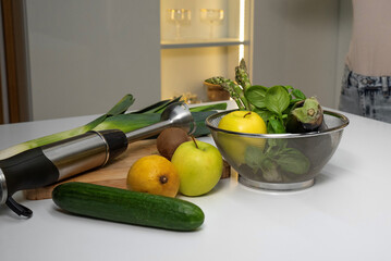 Immersion blender on kitchen table with green vegetables and fruits
