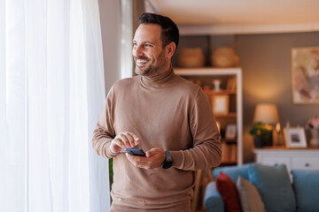 Happy mid adult man with mobile phone looking through window thoughtfully while standing in living room