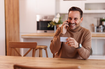 Smiling mid adult man with credit card talking over mobile phone while sitting at dining table