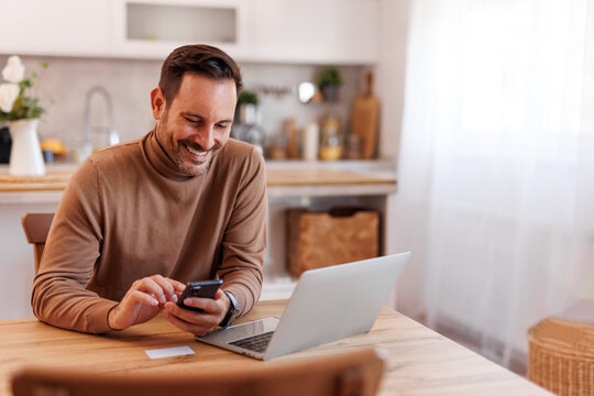 Portrait of smiling mid adult man with credit card using mobile phone and laptop on dining table at home