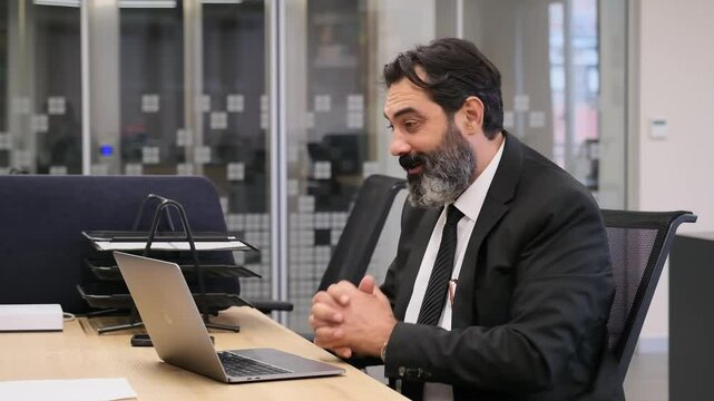 A middle-aged, dark-haired, bearded businessman is in  office at desk, clapping his hands during a video meeting on his computer to congratulate his employees