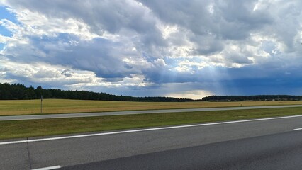 Fototapeta premium An asphalt road with markings is laid out among agricultural fields. A power line runs nearby, and behind it a forest grows and a village stands. On a summer day, sunlight breaks through thick clouds