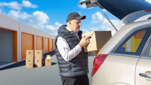 Man puts box in trunk car. Tenant of storage unit. Guy takes things from his personal storage. Man with car near street warehouse units. Guy unloads goods for safekeeping. Pantrys with orange gates
