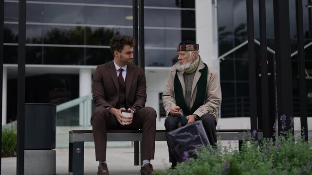 Friendly young businessman sitting on bench and giving takeaway coffee to homeless elderly man, showing compassion and generosity in urban environment. Concept of helping older tramp people.