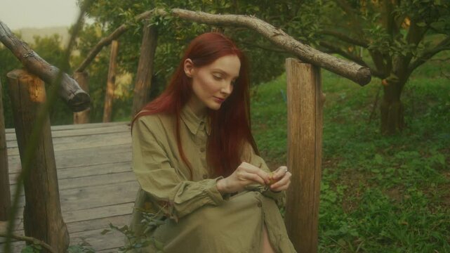 A ginger young woman sits on a wooden bridge in an orange orchard peeling an orange