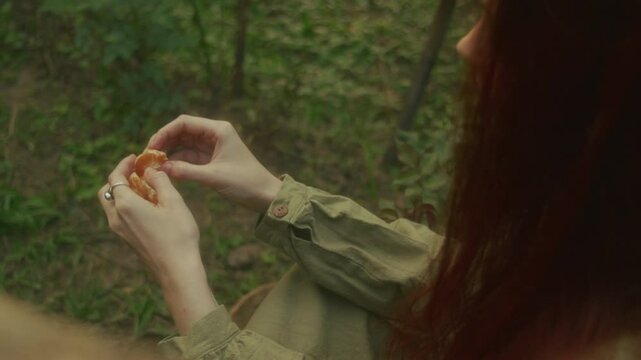 A ginger young woman sits on a wooden bridge in an orange orchard peeling an orange