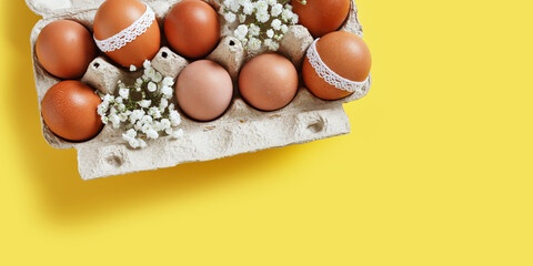 Brown eggs decorated with lace and white gypsophila flowers in carton on yellow background, banner. Easter celebration concept. Above photo with festive food. Top view of painted chicken egg minimal