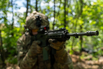 A soldier in full camouflage gear takes aim with a rifle in a wooded area, showcasing military equipment and focus in a natural setting. military operations in the forest