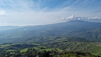 Expansive rural landscape with hills, greenery, and distant mountains under a partly cloudy sky.
