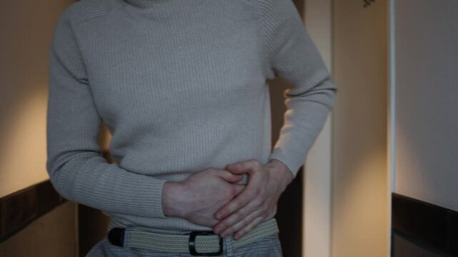 A person in a beige turtleneck and plaid trousers stands in a hallway, holding their side with visible discomfort, suggesting abdominal pain. The soft lighting emphasizes a quiet, introspective atmosp
