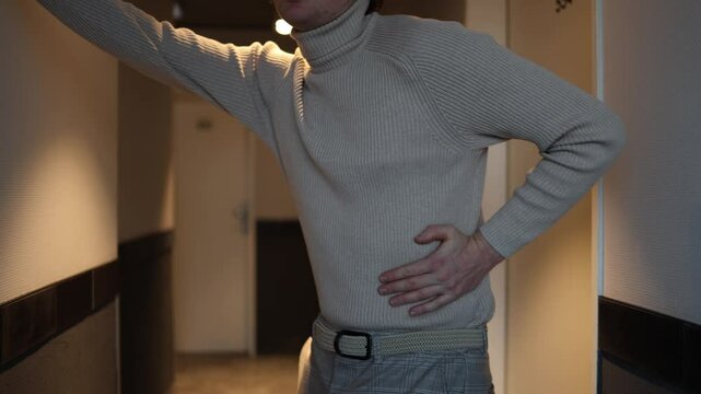 A person in a beige turtleneck and plaid trousers is standing in a hallway, holding their lower back as if experiencing discomfort. The warm lighting adds an introspective, moody atmosphere.





