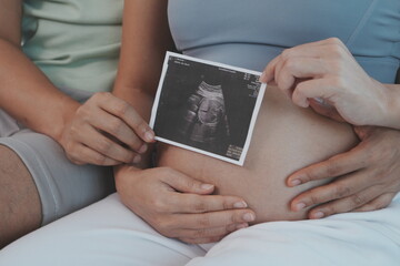 Asian husband and wife sit together hugging each other, father holds wife's seven-month pregnant belly and both are happy to have ultrasound pictures.