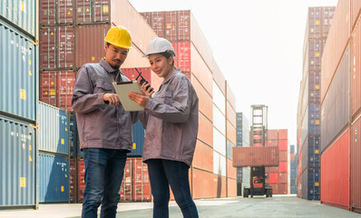 Two workers standing in cargo shipping container yard inspect documents on digital tablet. Male and female engineer wearing uniform and hard hats using walkie talkie for manage container loading. Tra