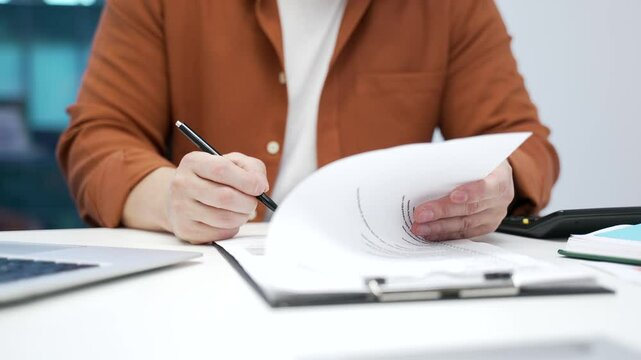 Close up of a man's hand signing documents at a desk at a workplace in a business office. Asian boss businessman executive looks through the folder with documentation and signs the contract with a pen