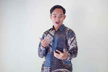 Shocked young Asian man wearing batik shirt looking at smartphone with surprised facial expression isolated on white background