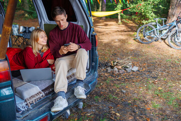 A young couple while relaxing in the forest in a homemade camper van has fun watching something on a smartphone and laptop