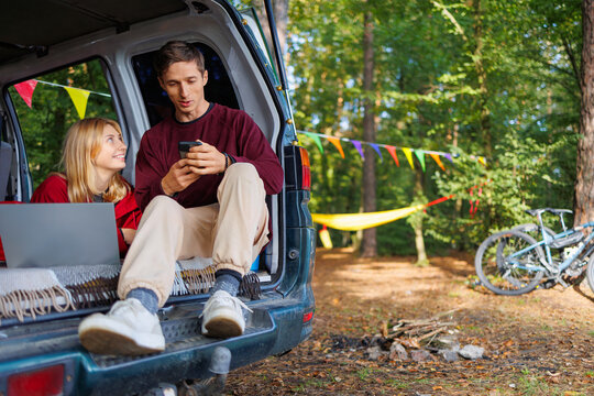 A young couple while relaxing in the forest in a homemade camper van has fun watching something on a smartphone and laptop