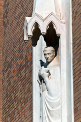 A statue on Szeged's Votive Church shows a monk holding a fish, with a live bird perched on it. The Gothic-inspired sculpture, set against a brick wall, captures a tranquil moment.