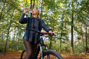 A young woman on a bicycle in the forest drinks water from a bottle, restoring her water balance after riding
