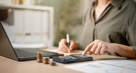 calculator, balance, accounting, analyzing, finance, wealth, audit, chart, graph, profit. A woman is sitting with a calculator. She is writing on a paper and pointing at a number on the calculator.