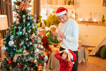 merry christmas, party, celebration, celebrate, present, childhood, christmas, parents, parent, parenthood. A man is standing in front of a Christmas tree. The scene is festive and joyful.