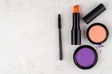 A vibrant makeup flat lay featuring purple eyeshadow, orange lipstick, cream foundation, and black mascara