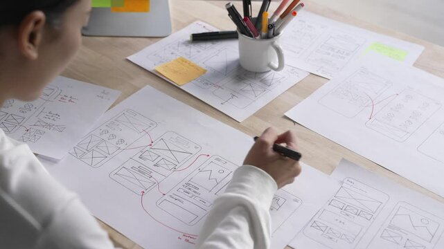 UX UI designer working on wireframes for a website and mobile app prototype, surrounded by sketches of user-flow and design tools, in concept of website and mobile application design concept.