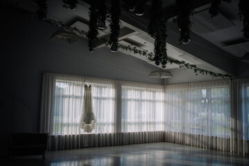 A bridal gown hangs in an empty, softly lit room with sheer curtains and greenery on the ceiling, creating an elegant and serene wedding preparation scene..