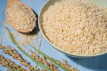 A bowl of brown rice is placed on a blue table next to a spoonful of raw rice and ears of rice.A bowl is filled with rice and the spoon is full.The rice is spread out in the bowl.