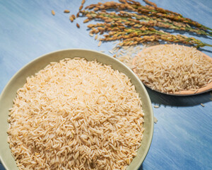 A bowl of rice is placed on a blue table next to a spoonful of rice and ears of rice.The bowl is filled with rice and the spoon is full.The rice is spread out in the bowl.