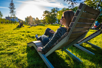 Beautiful middle aged woman wearing jeans, blouse, sneakers and sunglasses sitting on deck chair in city park on sunny spring day. Side view