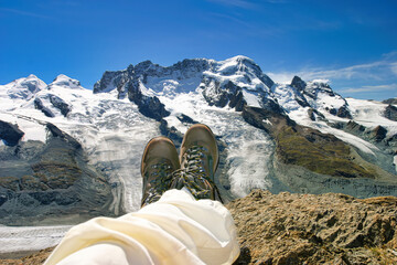 Wandern in den Schweizer Alpen mit Blick auf verschneite Berge. Eine Person sitzt mit Bergstiefeln auf einem Felsen und genießt den atemberaubenden Blick auf schneebedeckte Gipfel und einen Gletscher 