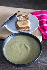 Broccoli soup puree in a black ceramic bowl with grilled cheese sandwich on a white ceramic plate on a wooden rustic dining table with red tea towel