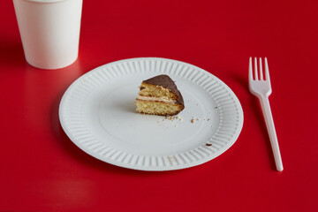 A small piece of chocolate pie on a white disposable plate and a white paper cup nearby against a red background, a portion of dessert, diet, small serving.