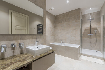A spacious bathroom decorated with beige tiles, with a glass shower and a large mirror .over the sink.