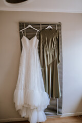 Two dresses hang side by side on personalized hangers, one white bridal gown and the other an elegant green dress, displayed indoors against shutters..