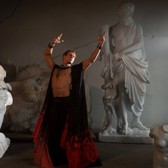 Fototapeta premium Muscular charismatic bodybuilder model in a silk cape makes an expressive pose, sculptor's workshop