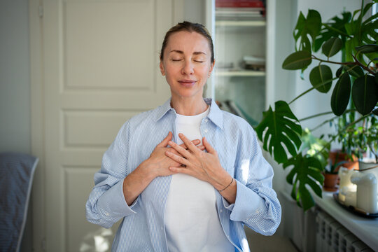 Calm peaceful woman standing with closed eyes, folding one's arms on chest meditating and doing breathing exercises. Mental balance recovery, slowly life, doing rituals to restore mental state.