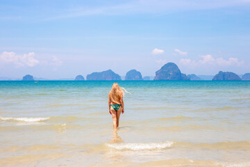 A woman with long blond hair in a swimsuit walks into the sea, seen from behind, with towering mountains in the background.