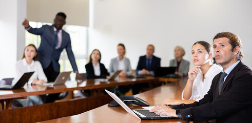 Side portrait of mid aged white dark-haired male manager attending business meeting in conference room and interestedly watching colleague's presentation together with young white female coworker