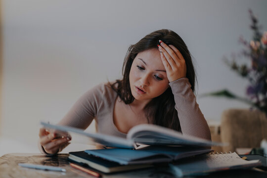 A young college student appears stressed while studying at home, surrounded by textbooks and notes. She is concentrating hard, holding her head, signifying the challenges of academic life.