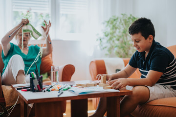 A grandmother is knitting a vest as her young nephew enjoys playing with building blocks on the table. The cozy home setting reflects warmth and family bonding.