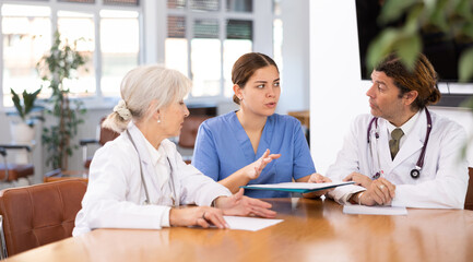 Fototapeta premium Focused confident young female doctor conducting medical council with experienced colleagues sitting at table with papers in office
