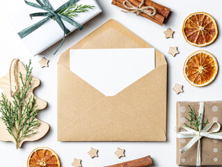 Envelope with a blank card decorated with a small sprig of greenery, surrounded by gift boxes wrapped with twine, pine cones, dried orange slices, and wooden ornaments on a light background. 