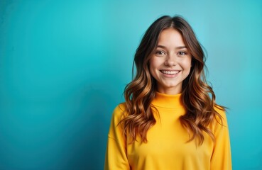Fototapeta premium Young woman in yellow sweater smiles at camera. Happy female with brown hair poses against blue background. Pretty girl is looking at viewer, portrait for fashion, beauty blog, social media content.