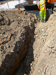 Construction worker and excavator performing trench excavation and site preparation for foundation digging. Ground compaction, soil stabilization, and erosion control on construction site.